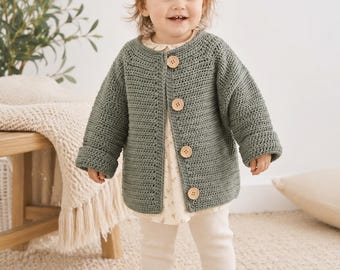 May include: A sage green crochet cardigan with three wooden buttons. The cardigan is worn over a floral print dress and cream-colored leggings. The outfit is completed with matching shoes. The child is smiling and wearing a white bow.
