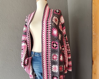 May include: A long, gray crochet cardigan with pink and white accents. The cardigan has a fringe trim and is displayed on a mannequin.