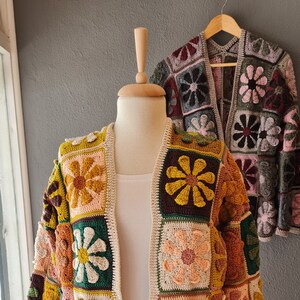May include: A crocheted cardigan with a floral pattern in shades of brown, green, yellow, and pink. The cardigan is open in the front and has a relaxed fit.