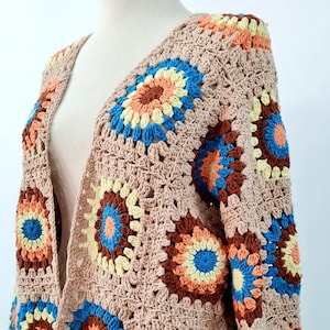 Puede incluir: Un cárdigan de ganchillo con un frente largo y abierto y mangas largas. El cárdigan está hecho de hilo beige y presenta un patrón geométrico colorido con tonos de azul, naranja, marrón y amarillo.
