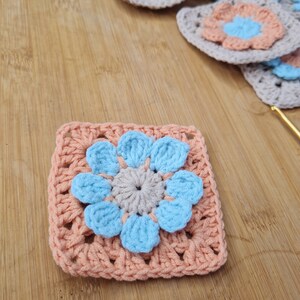 May include: A crocheted square with a light blue flower in the center. The flower has a light gray center and is surrounded by peach-colored crocheted stitches.
