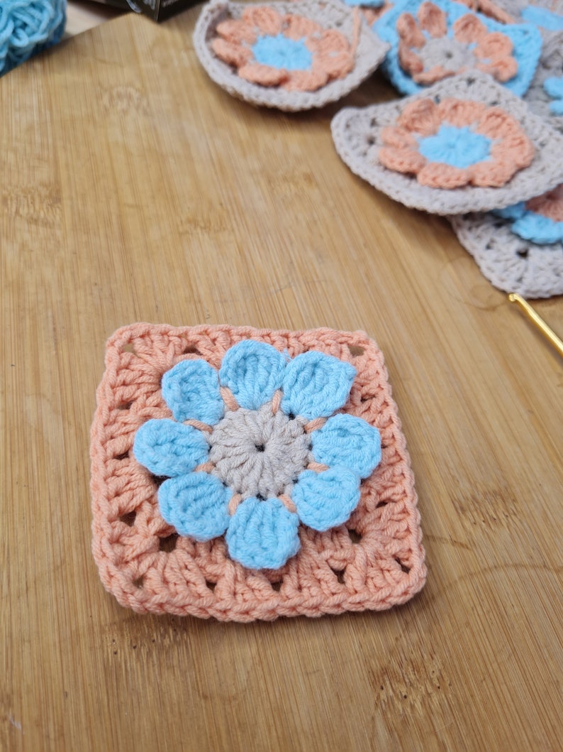 May include: A crocheted square with a light blue flower in the center. The flower has a light gray center and is surrounded by peach-colored crocheted stitches.