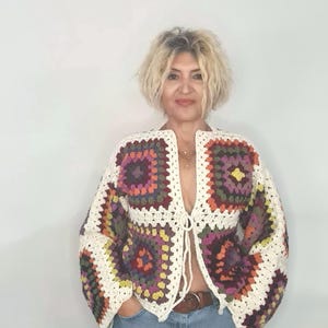 May include: A white crochet cardigan with a colorful geometric pattern. The cardigan has a tie closure at the waist and is worn over a pair of light blue jeans.