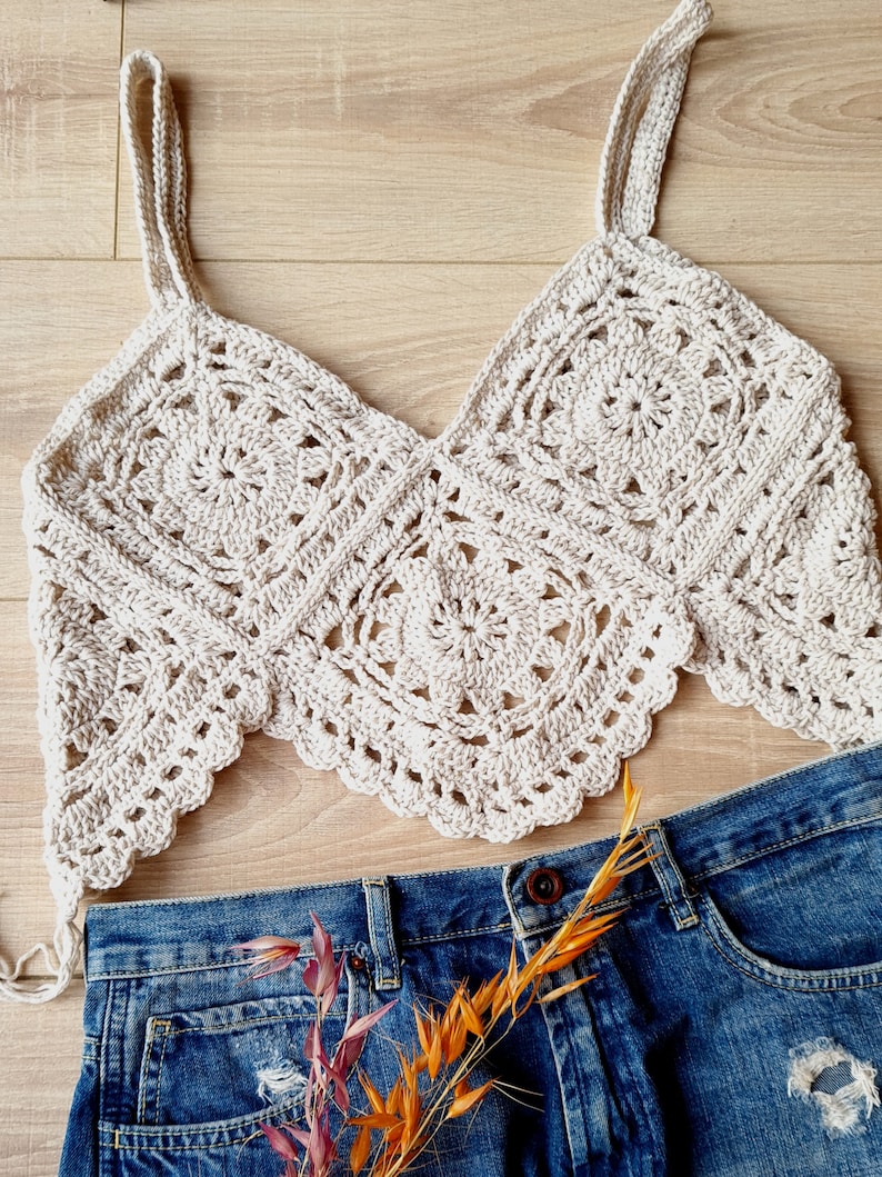 May include: A crocheted cream-colored top with a square pattern and thin straps. The top is laying on a wooden surface with a pair of blue jeans underneath.