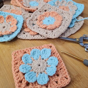 May include: A crocheted granny square with a light blue flower in the center. The flower has five petals and is surrounded by a peach-colored border. The square is made with a beige yarn.
