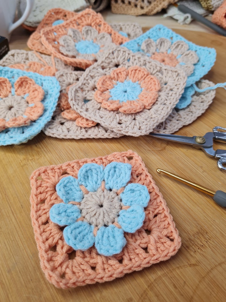 May include: A crocheted granny square with a light blue flower in the center. The flower has five petals and is surrounded by a peach-colored border. The square is made with a beige yarn.