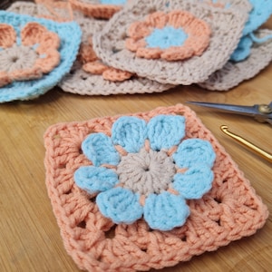 May include: A crocheted granny square with a peach colored border and a blue flower in the center. The flower has five petals and a smaller beige circle in the center.