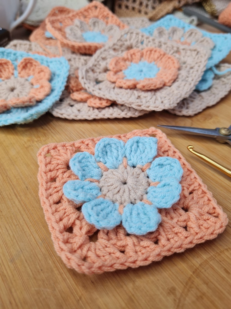 May include: A crocheted granny square with a peach colored border and a blue flower in the center. The flower has five petals and a smaller beige circle in the center.