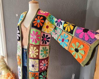 May include: A long, open-front crochet cardigan with a colorful floral pattern. The cardigan features square panels with various flower designs in shades of pink, orange, yellow, blue, and green. The sleeves are wide and bell-shaped, with a green border.