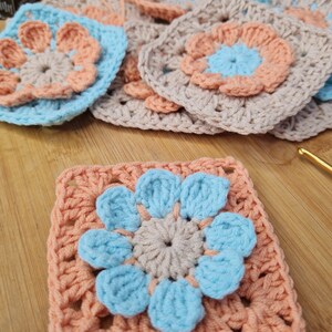 May include: A crocheted granny square with a blue and peach flower design. The center of the flower is a light brown circle.