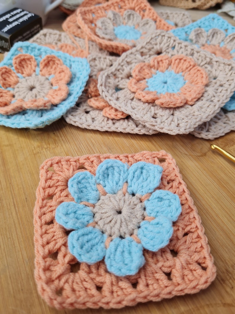May include: A crocheted granny square with a blue and peach flower design. The center of the flower is a light brown circle.
