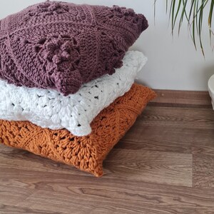 May include: Three crocheted throw pillows stacked on a wooden floor. The top pillow is a deep purple, the middle pillow is white, and the bottom pillow is a burnt orange.