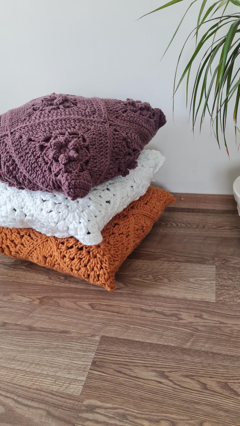 May include: Three crocheted throw pillows stacked on a wooden floor. The top pillow is a deep purple, the middle pillow is white, and the bottom pillow is a burnt orange.