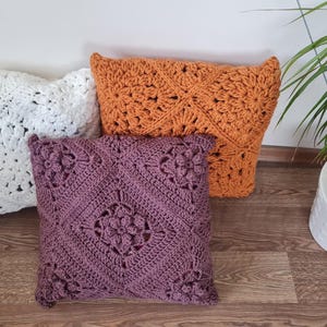 May include: Three crocheted throw pillows in different colors and patterns. The pillows are white, orange, and purple. The pillows are arranged on a wooden floor.