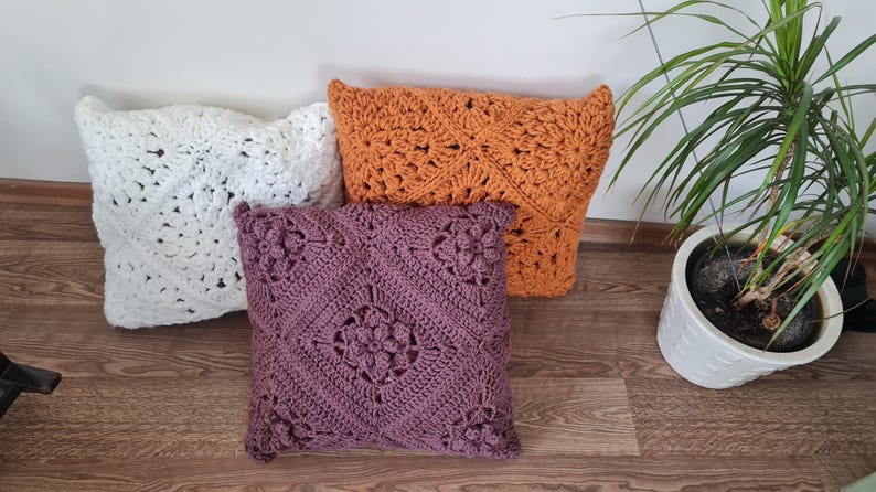 May include: Three crocheted throw pillows in different colors and patterns. The pillows are white, orange, and purple. The pillows are arranged on a wooden floor.