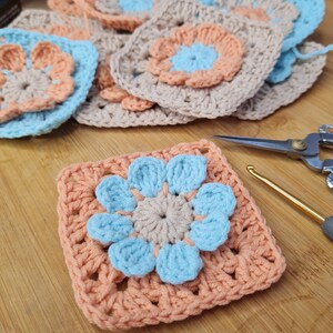 Op de afbeelding: Een close-up van een gehaakt granny square met een lichtblauwe bloem in het midden. De bloem heeft een lichtbruin midden en is omgeven door een perzikkleurige rand. Het vierkant ligt op een houten oppervlak.