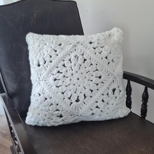 May include: White crocheted pillow with a square design featuring a large flower in the center. The pillow is sitting on a brown leather rocking chair.