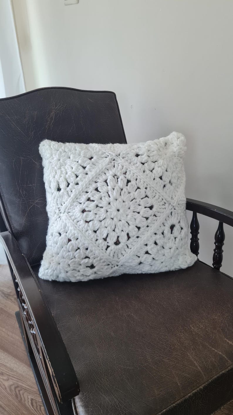 May include: White crocheted pillow with a square design featuring a large flower in the center. The pillow is sitting on a brown leather rocking chair.