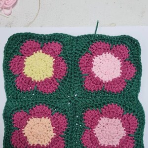 May include: A crocheted square with four flower motifs. The flowers are in shades of pink, yellow, and peach, and are surrounded by a green border.