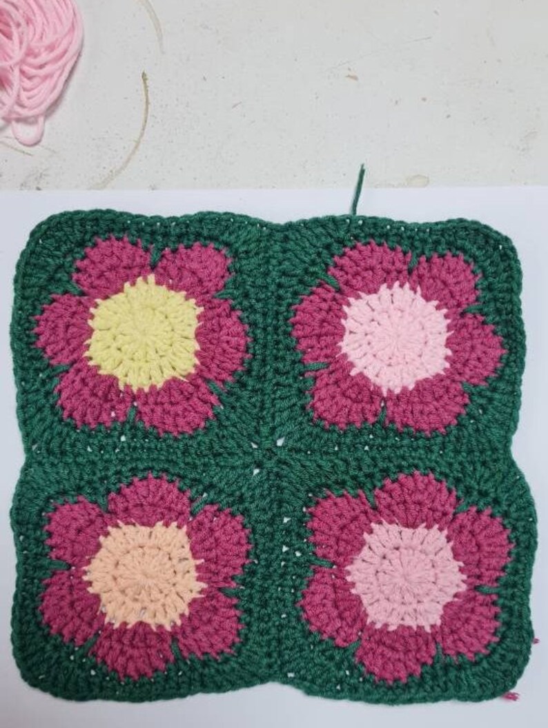May include: A crocheted square with four flower motifs. The flowers are in shades of pink, yellow, and peach, and are surrounded by a green border.
