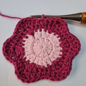 May include: A pink and burgundy crocheted flower with a textured design. The flower is made with yarn and has a circular shape.
