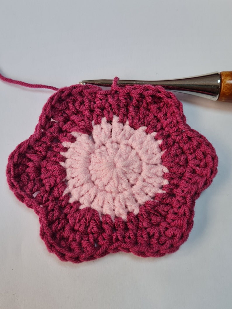 May include: A pink and burgundy crocheted flower with a textured design. The flower is made with yarn and has a circular shape.