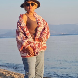May include: A woman wearing a handmade crochet cardigan with a floral pattern in shades of red and white. She is also wearing a brown tank top, blue jeans, a dark hat, and sunglasses. The background shows a beach and the ocean.