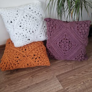 May include: Three crocheted throw pillows in different colors and patterns. The pillows are white, orange, and purple. The pillows are on a wooden floor.
