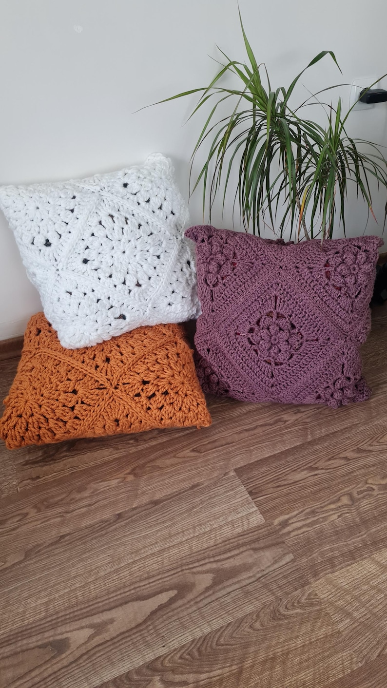 May include: Three crocheted throw pillows in different colors and patterns. The pillows are white, orange, and purple. The pillows are on a wooden floor.