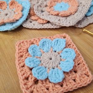 May include: A crocheted square with a light blue flower in the center. The flower has five petals and a smaller, beige center. The square is surrounded by a border of peach-colored yarn.