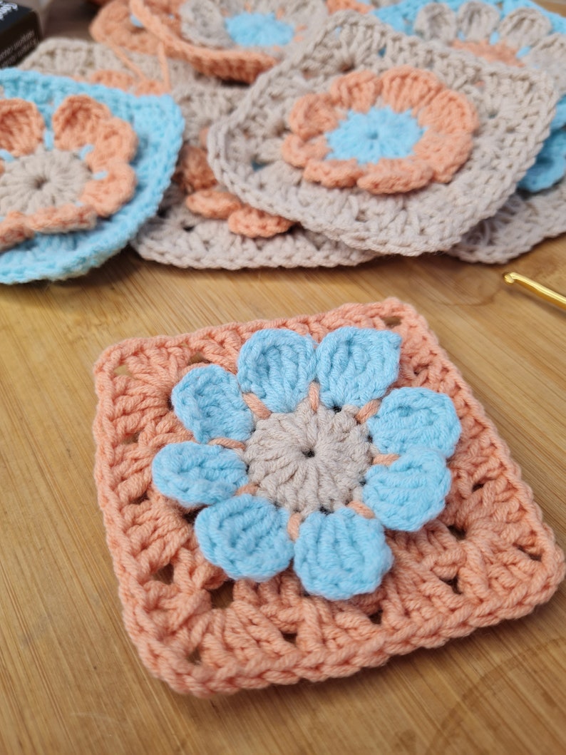 May include: A crocheted square with a light blue flower in the center. The flower has five petals and a smaller, beige center. The square is surrounded by a border of peach-colored yarn.