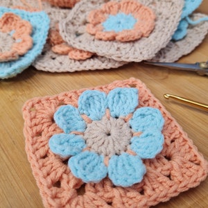 May include: A crocheted square with a peach colored border and a blue flower in the center. The flower has five petals and a smaller beige flower in the center.