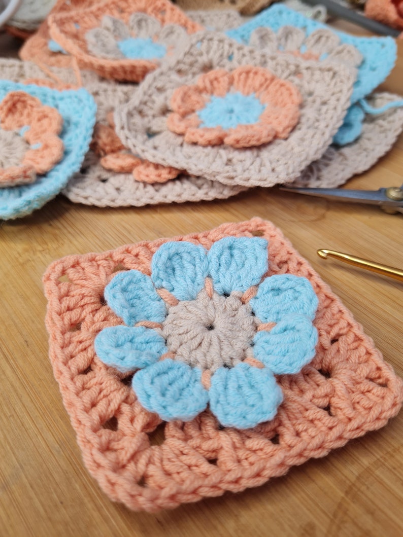 May include: A crocheted square with a peach colored border and a blue flower in the center. The flower has five petals and a smaller beige flower in the center.