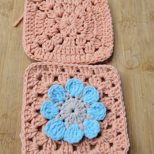 May include: Two crocheted squares in a peach color. The top square has a simple design. The bottom square has a blue flower in the center.
