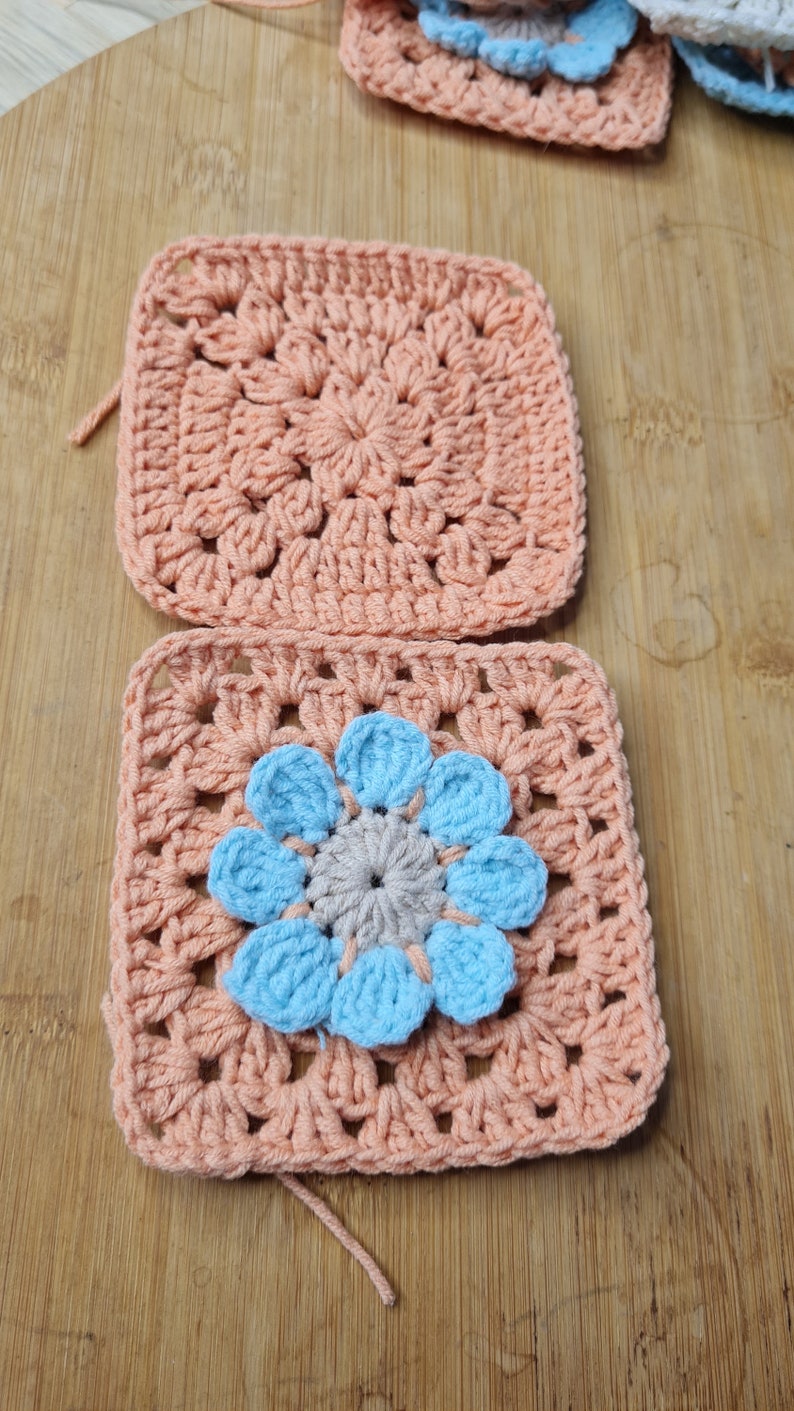 May include: Two crocheted squares in a peach color. The top square has a simple design. The bottom square has a blue flower in the center.