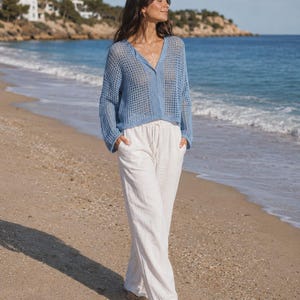 May include: A light blue, open-weave knit cardigan with long sleeves and a V-neck, paired with white linen pants. The outfit is worn on a sandy beach with the ocean in the background. The cardigan is a relaxed fit.