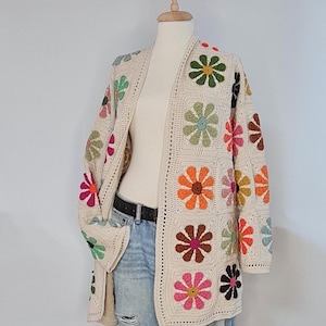 May include: Cream-colored crochet cardigan with colorful flower motifs. The open-front sweater features a square pattern with various flower designs in shades of pink, orange, green, and brown. The cardigan is displayed on a mannequin with blue jeans.
