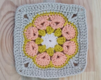 May include: A crocheted square with a flower design in shades of peach, yellow, and white. The square has a border of gray crochet.