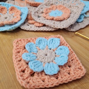 May include: A crocheted square with a peach background and a blue flower in the center. The flower has five petals and a smaller peach circle in the center.