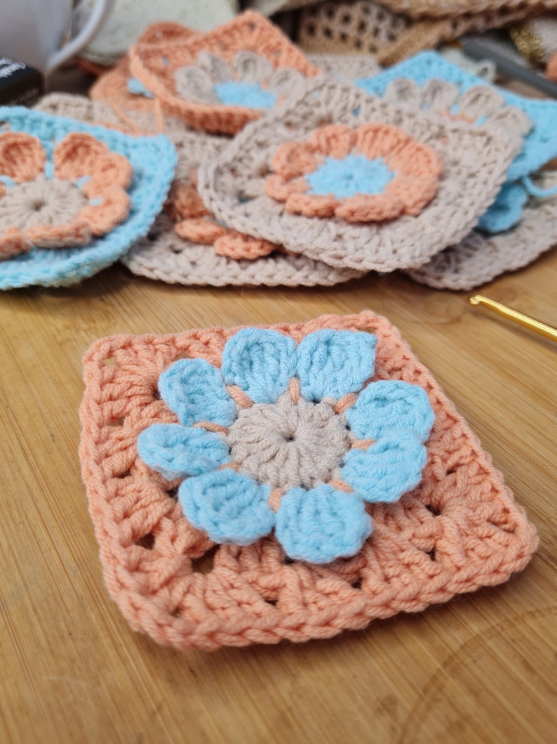 May include: A crocheted square with a peach background and a blue flower in the center. The flower has five petals and a smaller peach circle in the center.