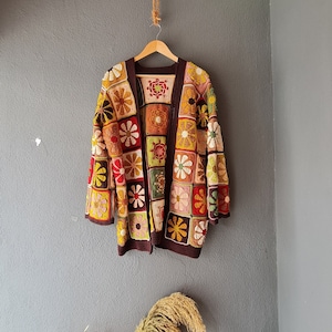 May include: A brown crocheted cardigan with a patchwork design featuring colourful flowers. The cardigan has a square shape and is made of a soft, textured yarn.