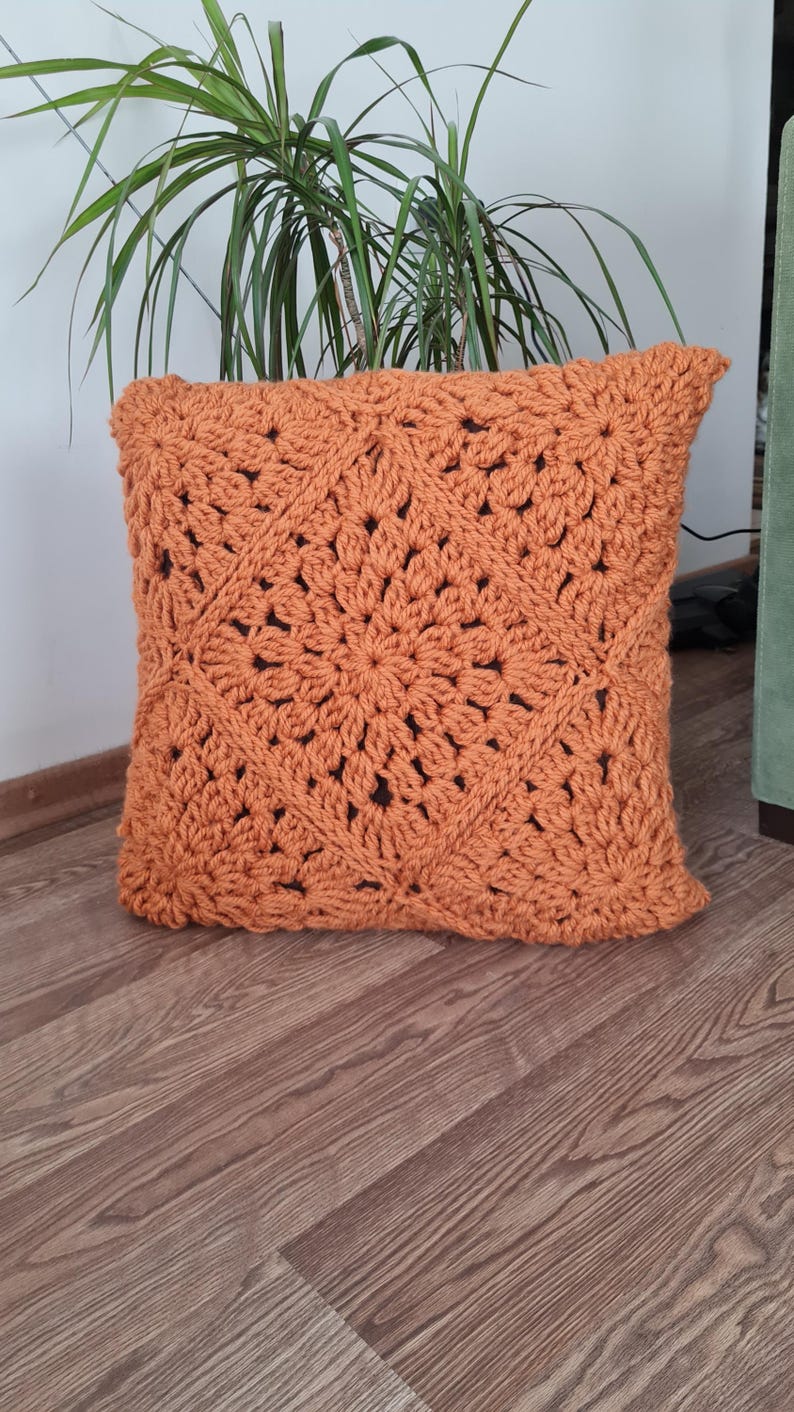 May include: A crocheted pillow with a square pattern in a burnt orange color. The pillow is made with a thick yarn and has a textured surface.