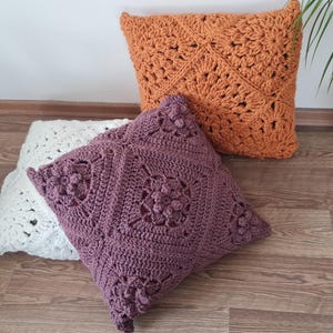 May include: Three crocheted throw pillows in different colors and patterns. The pillows are on a wooden floor. The pillow on the left is white, the middle pillow is purple, and the pillow on the right is orange.