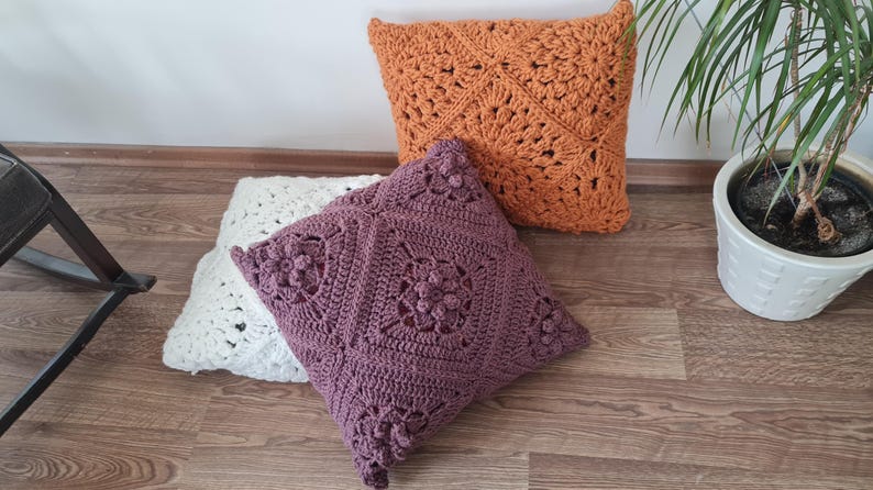 May include: Three crocheted throw pillows in different colors and patterns. The pillows are on a wooden floor. The pillow on the left is white, the middle pillow is purple, and the pillow on the right is orange.