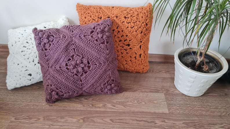 May include: Three crocheted throw pillows in different colors and patterns. The pillows are white, purple, and orange. They are arranged on a wooden floor.