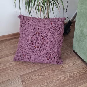 May include: A square, purple crocheted pillow with a floral design. The pillow is on a wooden floor.