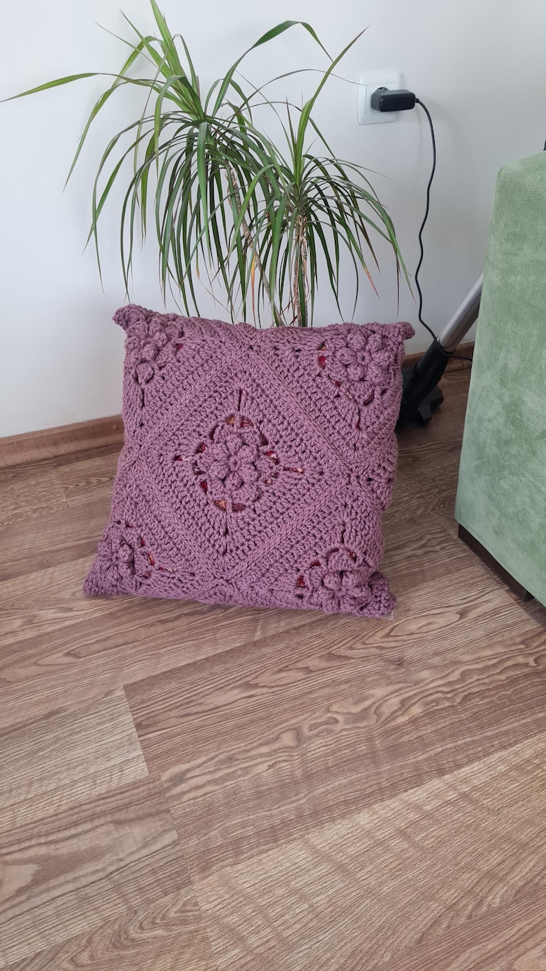 May include: A square, purple crocheted pillow with a floral design. The pillow is on a wooden floor.