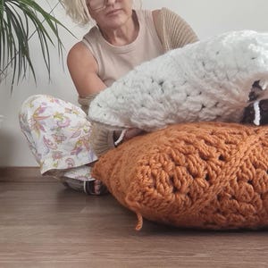 May include: A woman sits on a wooden floor holding two crocheted pillows. One pillow is white and the other is a burnt orange color.