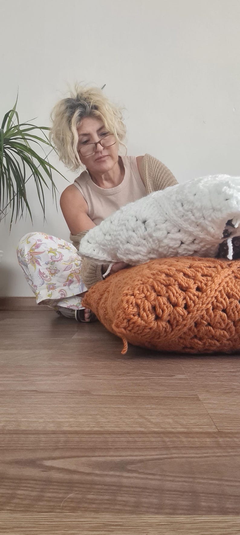 May include: A woman sits on a wooden floor holding two crocheted pillows. One pillow is white and the other is a burnt orange color.