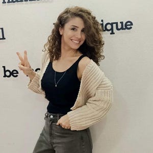 以下が含まれることがあります： 茶色の巻き毛の女性が、黒いタンクトップの上にベージュのニットカーディガンを着て、濃い緑色のジーンズを履いています。彼女は笑顔でピースサインをしています。背景は白い壁で、「humble」と「be unique」の文字が書かれています。
